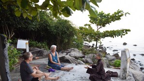 Three people sitting on mats outdoors near a body of water, with rocks and greenery in the background. - Island Smiles Homestay (Da Nang)