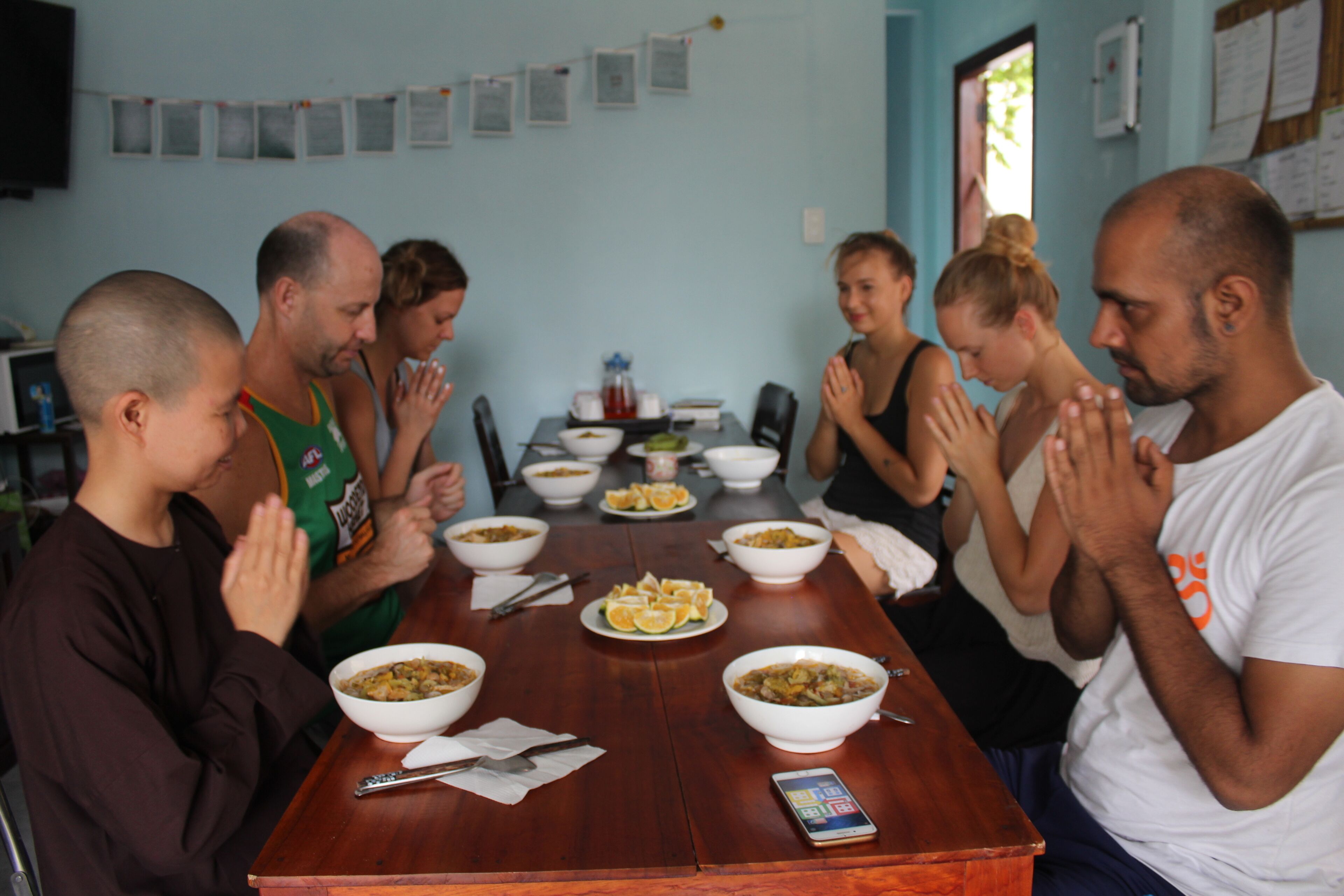 Un grupo de personas sentadas alrededor de una mesa de madera con platos de comida, en una habitación con paredes azul claro y un tablón de anuncios.