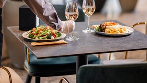 A dining table with two plates of food and two glasses of wine, set in a modern restaurant with large windows and potted plants. - Messier 53 Hotel Yerevan (Yerevan)
