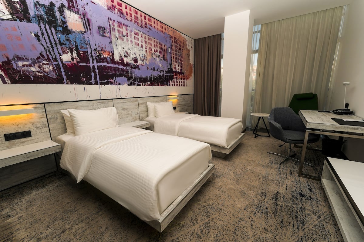 Een hotelkamer met twee bedden, een bureau, een stoel en een groot abstract schilderij aan de muur.