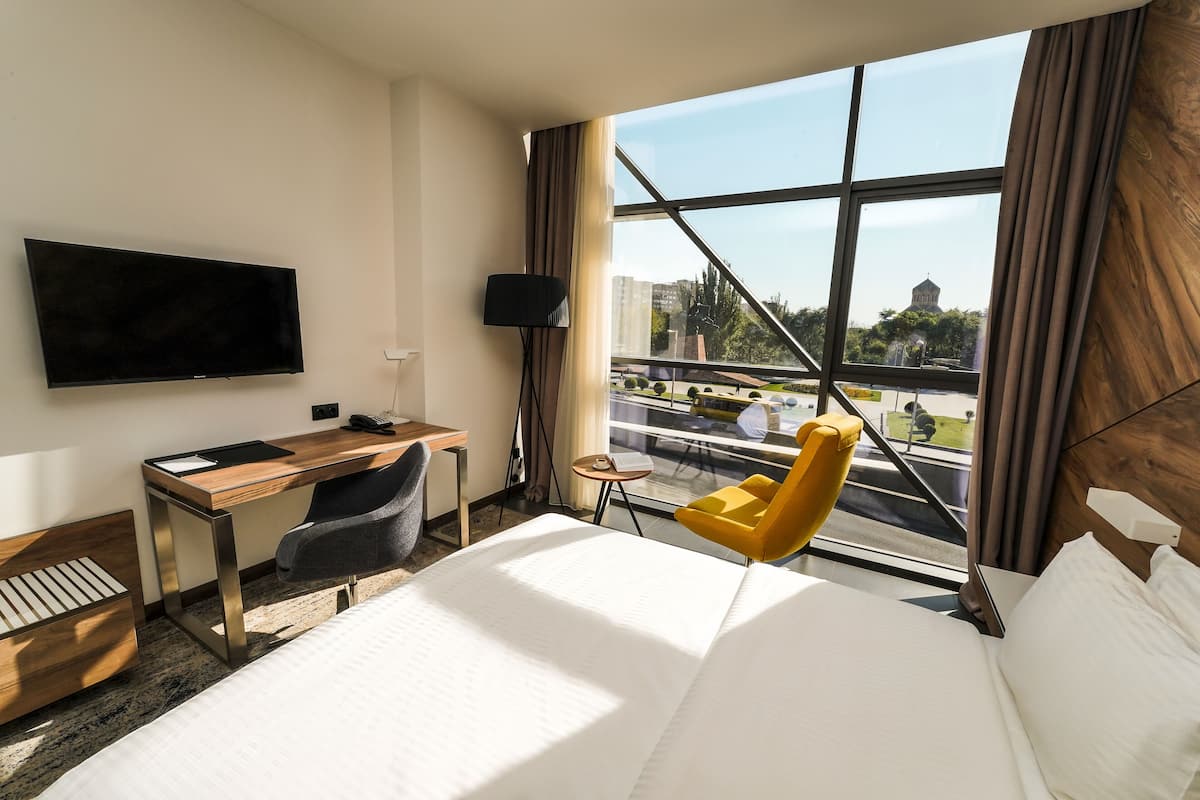Een hotelkamer met een bed, een bureau, een stoel, een televisie en een raam met uitzicht op gebouwen en groen.