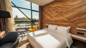 A modern hotel room with a large bed, a view of a cityscape, a wooden accent wall, and a small round table with a chair. - Messier 53 Hotel Yerevan (Yerevan)