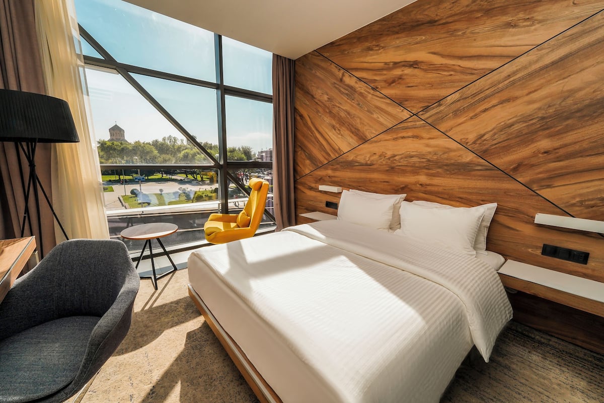 Een moderne hotelkamer met een groot bed, uitzicht op een stadsbeeld, een houten accentmuur en een klein rond tafeltje met een stoel.