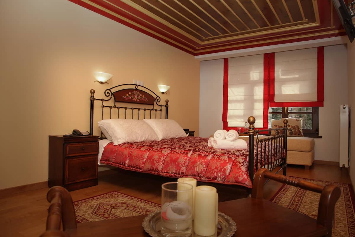 A bedroom with a bed, bedside tables, a dining table with chairs, a window with red curtains, and a view of the outdoors.
