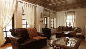 Un salon avec un canapé en cuir marron, une table basse en verre et un canapé beige. Il y a des rideaux aux fenêtres, un radiateur et une cheminée.