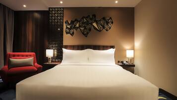 Habitación de hotel con una cama grande, un sillón rojo, dos mesitas de noche con lámparas y un panel decorativo en la pared.