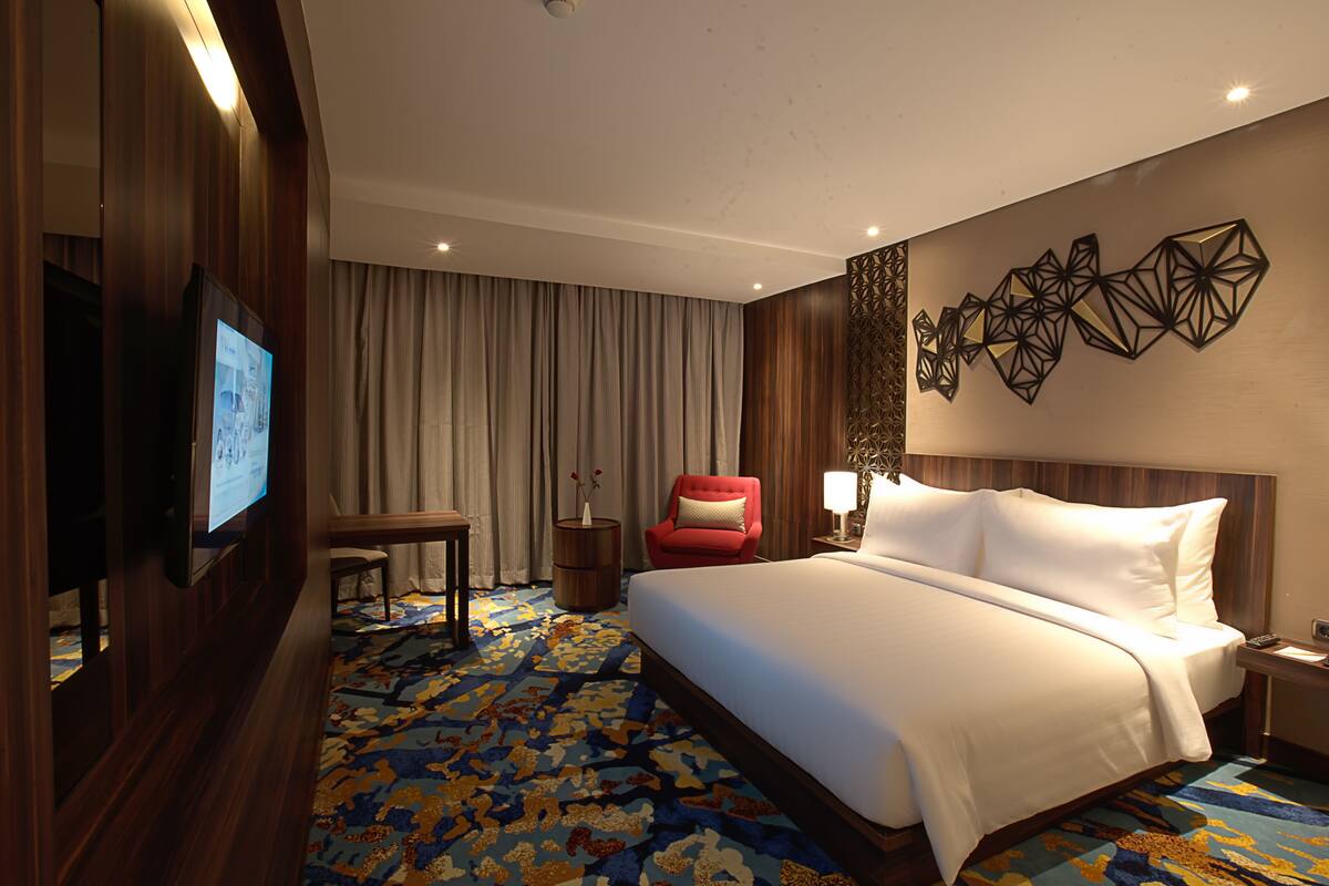 Een hotelkamer met een groot bed, een aan de muur bevestigde televisie, een rode fauteuil, een klein tafeltje en een decoratief wandobject.