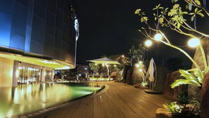 Vue nocturne d’un espace piscine d’hôtel, avec une terrasse en bois, une grande piscine, ainsi que des plantes et des parasols tout autour.