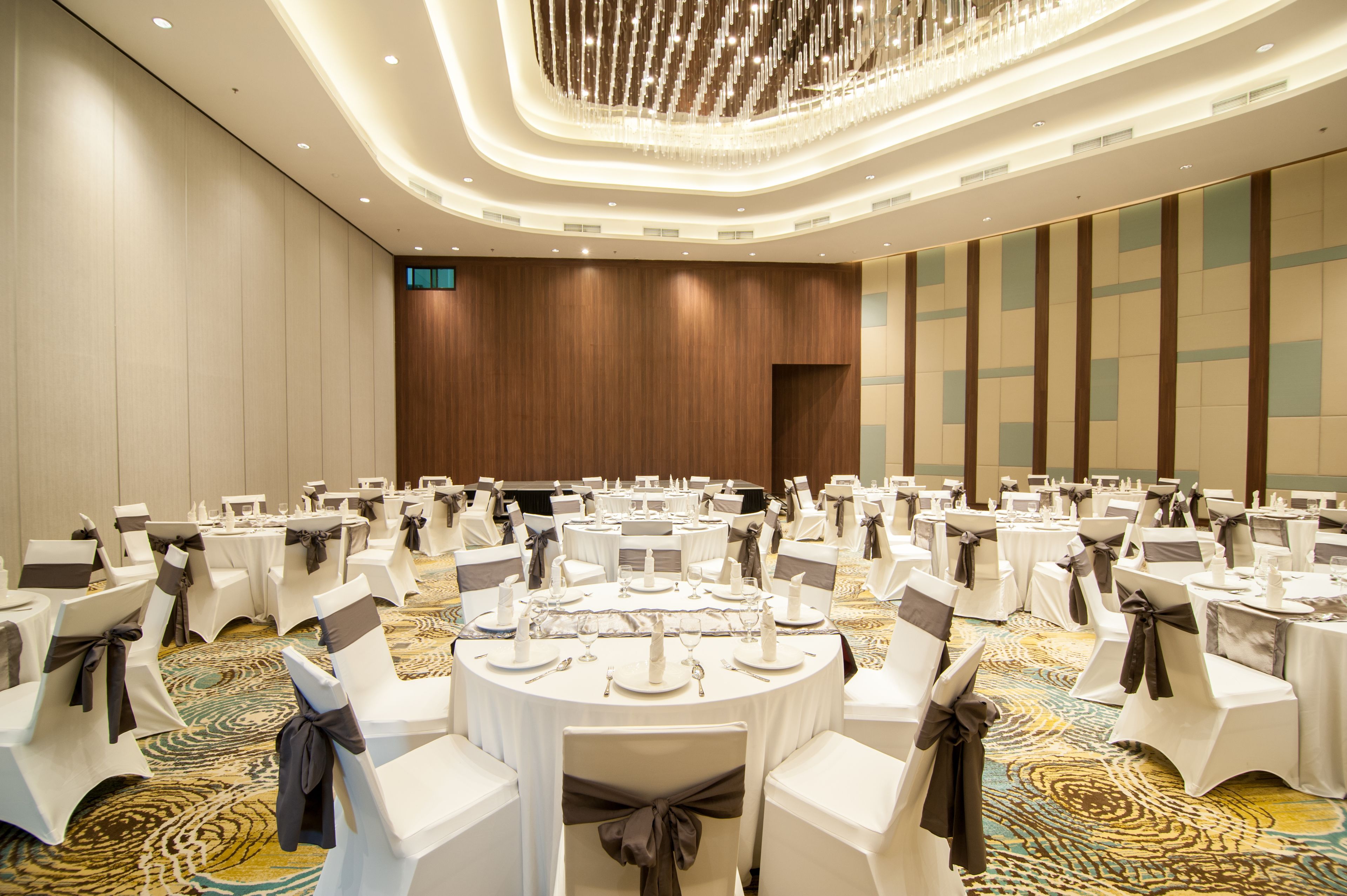 A banquet hall with round tables, white chairs with brown sashes, and a patterned carpet.