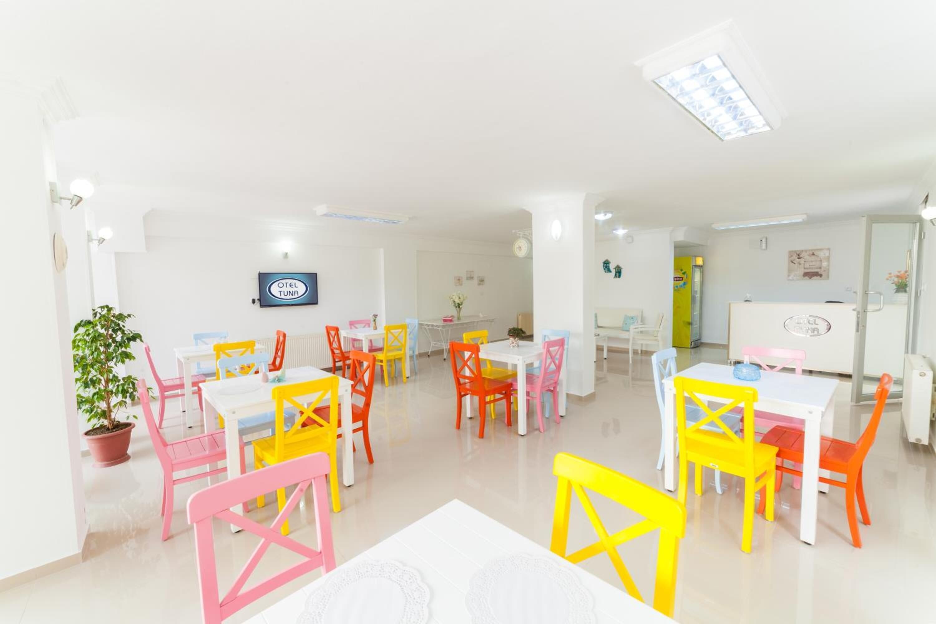 A brightly lit dining area with colorful chairs and tables, a potted plant, and a sign indicating the restaurant name.