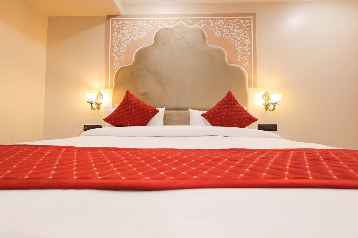 Hotelkamer met een groot bed, rood-witte beddengoed, twee rode kussens, een decoratief hoofdeinde en wandlampen.