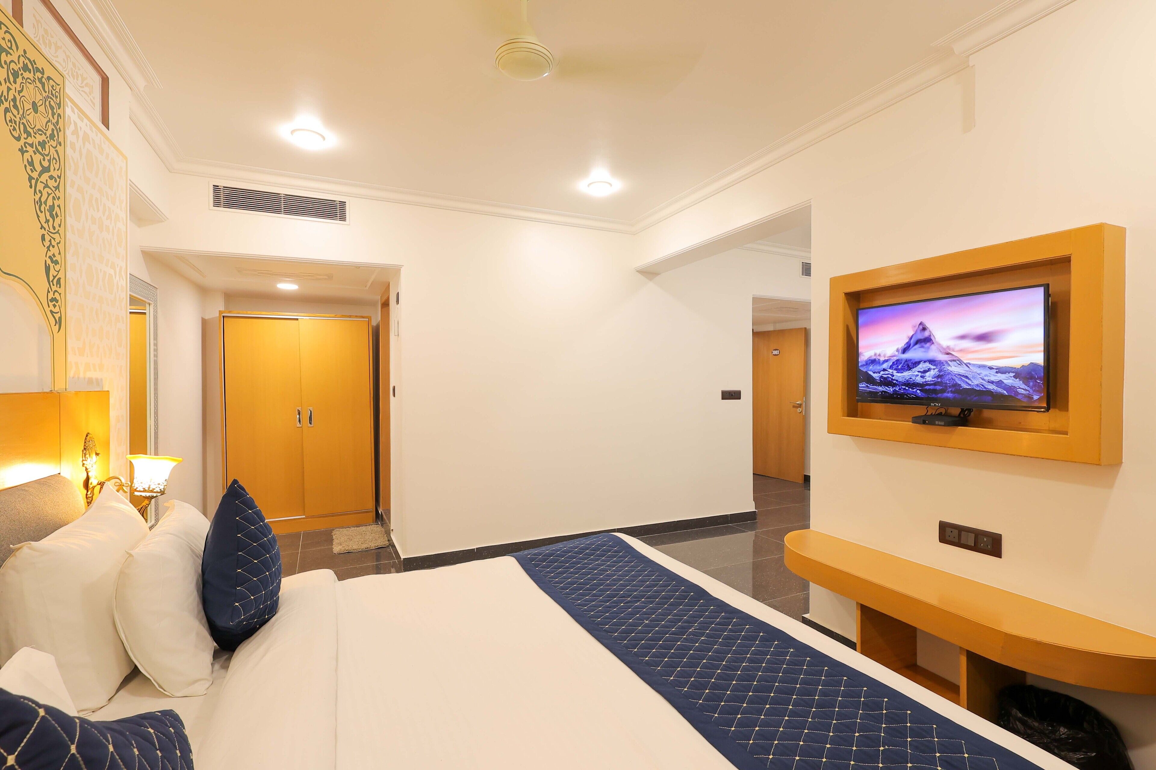 A hotel room with a bed, a TV mounted on the wall, a wooden door, and a hallway leading to another room.