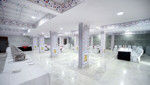 Un amplio salón de banquetes con una larga mesa dispuesta para una comida, rodeado de sillas blancas y un sofá.