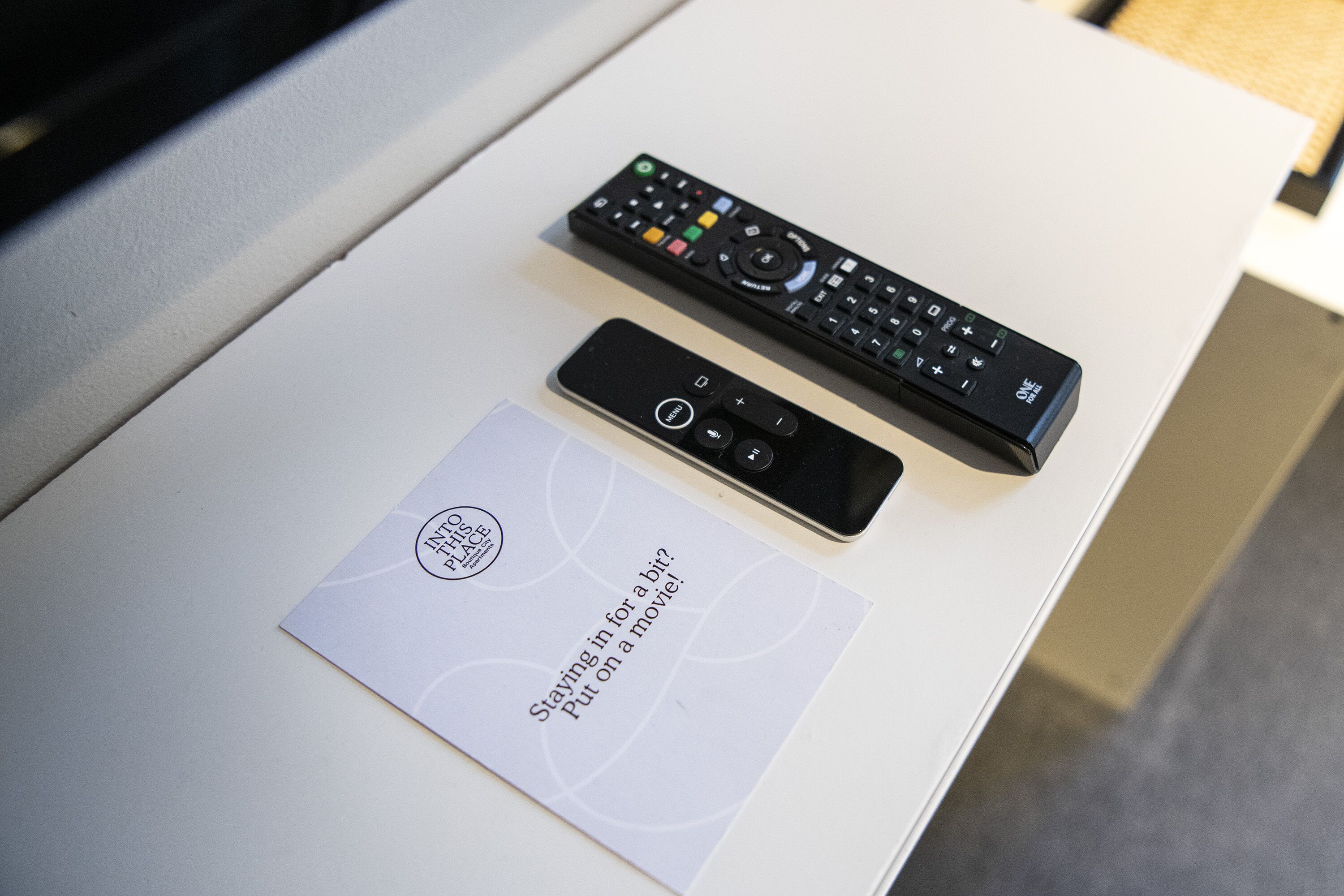 A remote control and a smartphone on a white table with a card that reads "Staying in for a bit. Put on a movie!"
