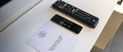 A remote control and a smartphone on a white table with a card that reads "Staying in for a bit. Put on a movie!"