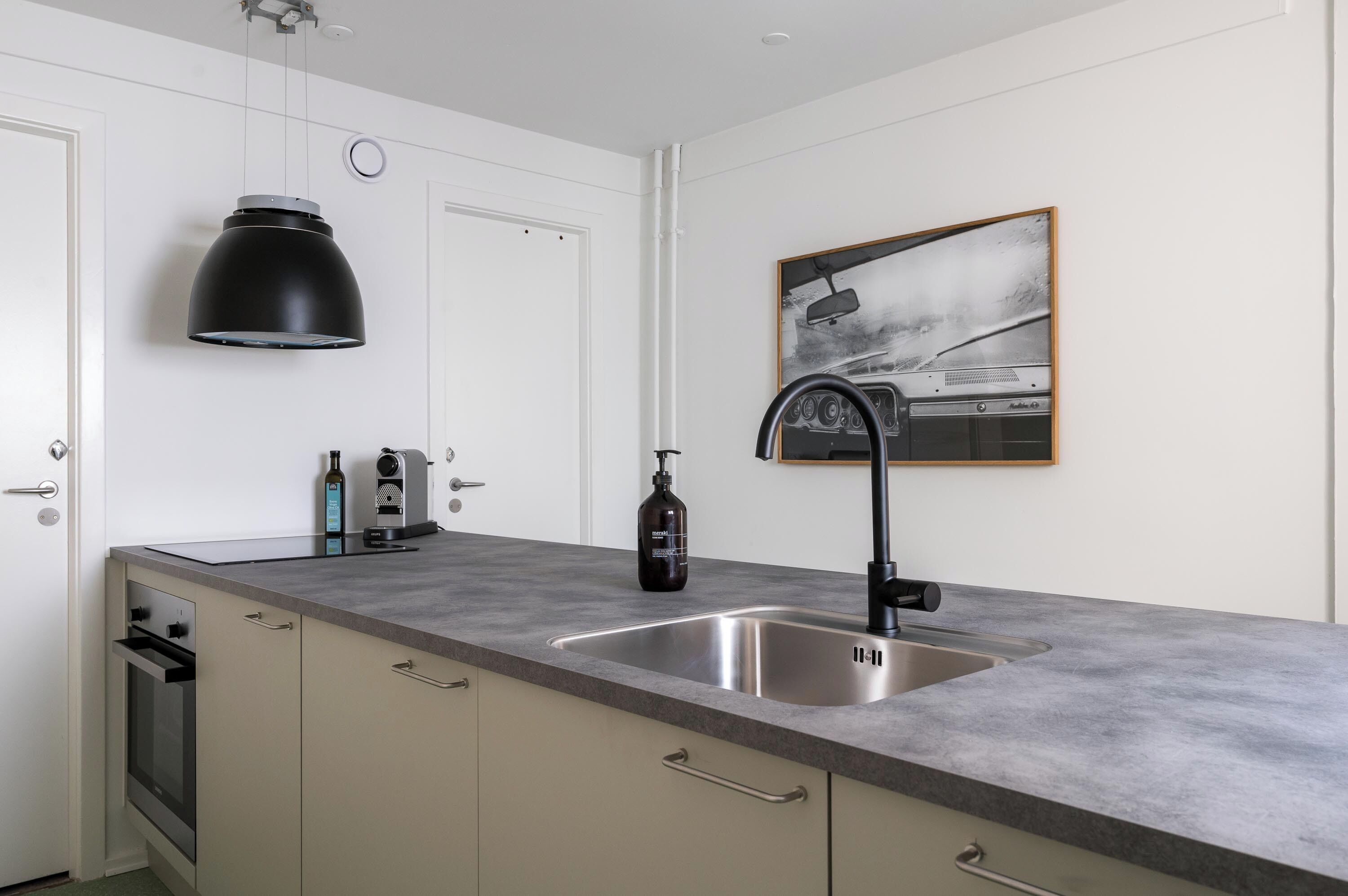 A modern kitchen with a grey countertop, stainless steel sink, and a black pendant light. There is a framed picture on the wall and a bottle of hand soap on the counter.