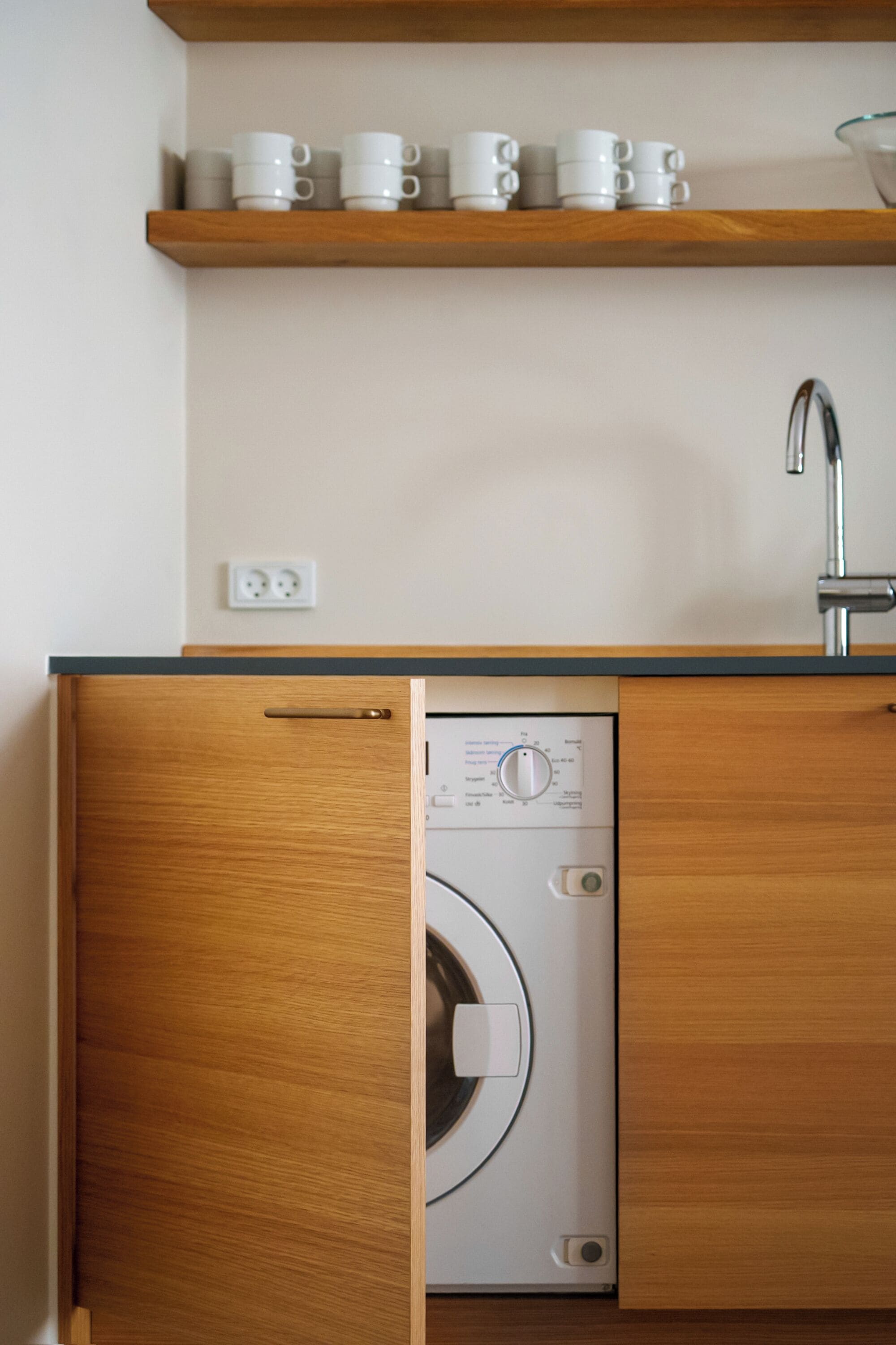Cocina compacta con lavadora integrada, cubierta de madera y estante con tazas blancas.