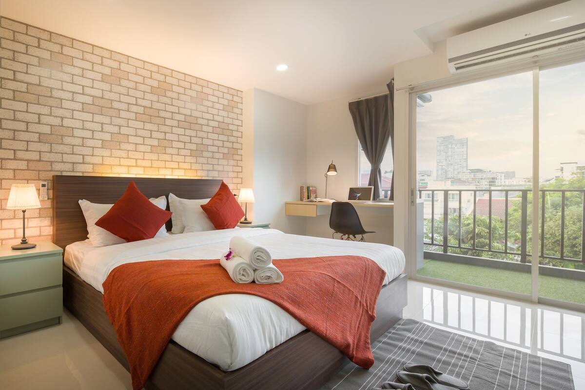 A modern hotel room with a brick wall, a large bed with red and white bedding, a desk with a laptop, and a balcony with a city view.