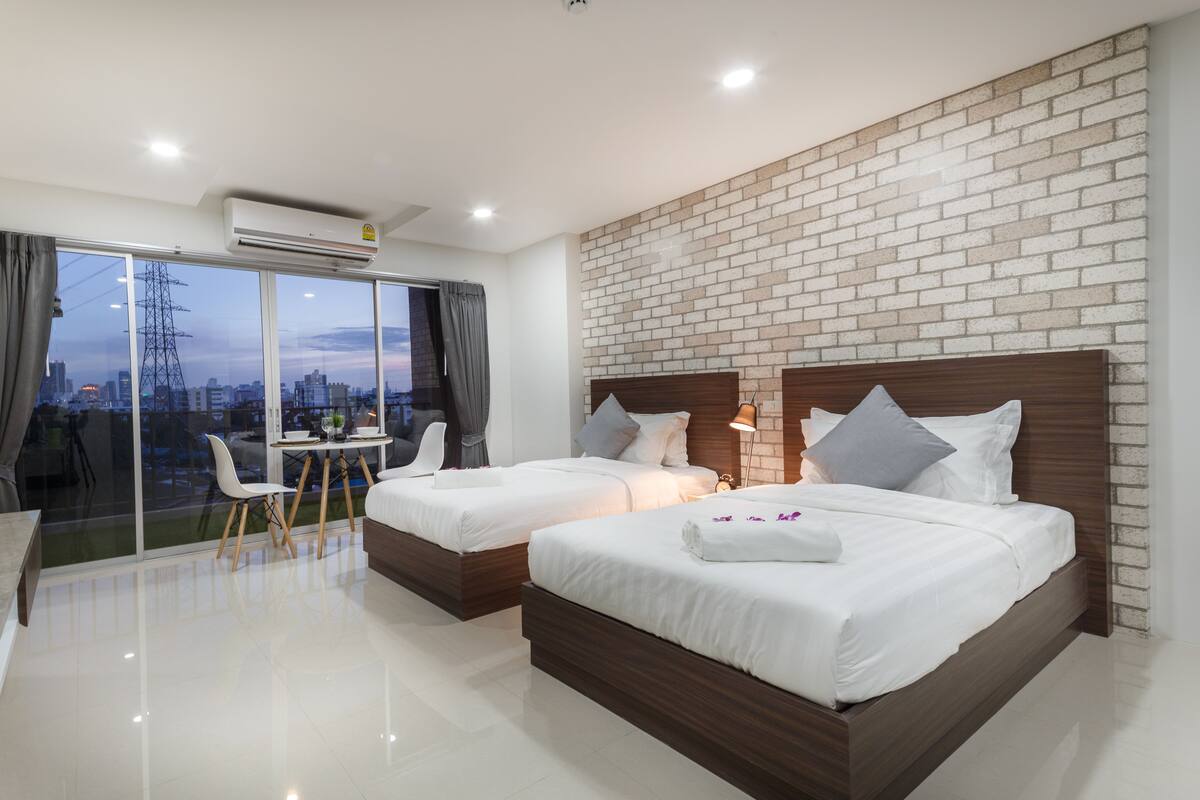 A modern hotel room with two beds, a dining area, and a large window with a city view.