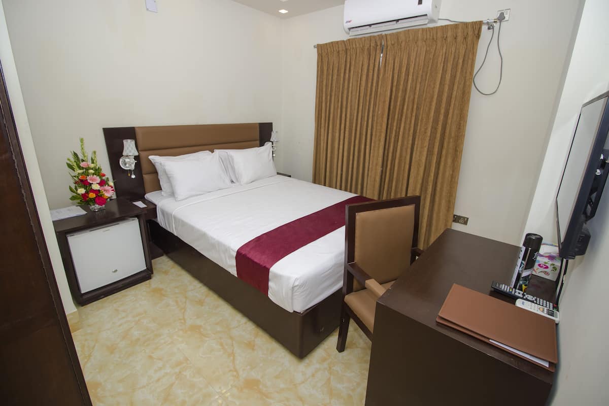 A hotel room with a bed, a desk, a chair, a small cabinet, and an air conditioning unit.