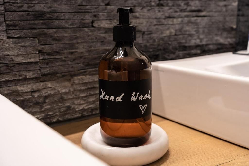 Un flacon de savon pour les mains est posé sur un porte-savon blanc dans une salle de bain dont un des murs est en pierre.