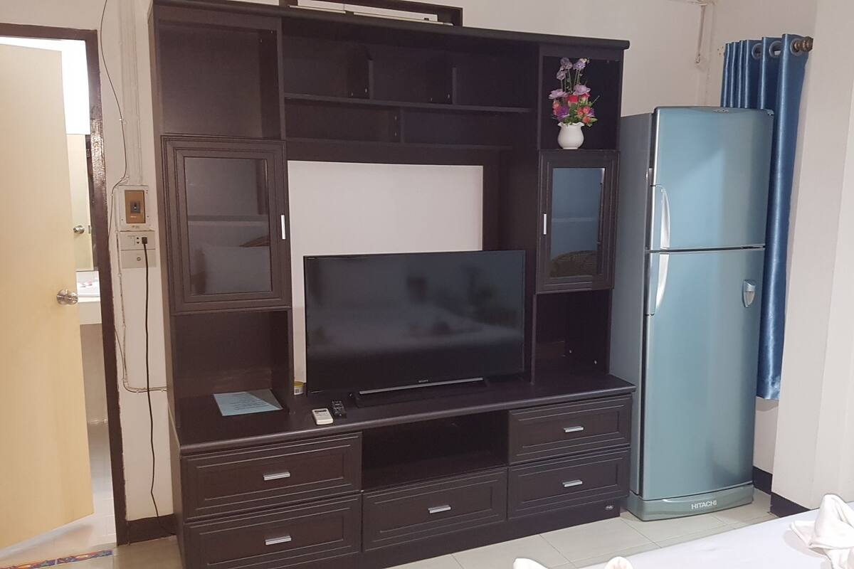 Un dormitorio con cama, televisión, refrigerador Hitachi y un mueble pequeño.