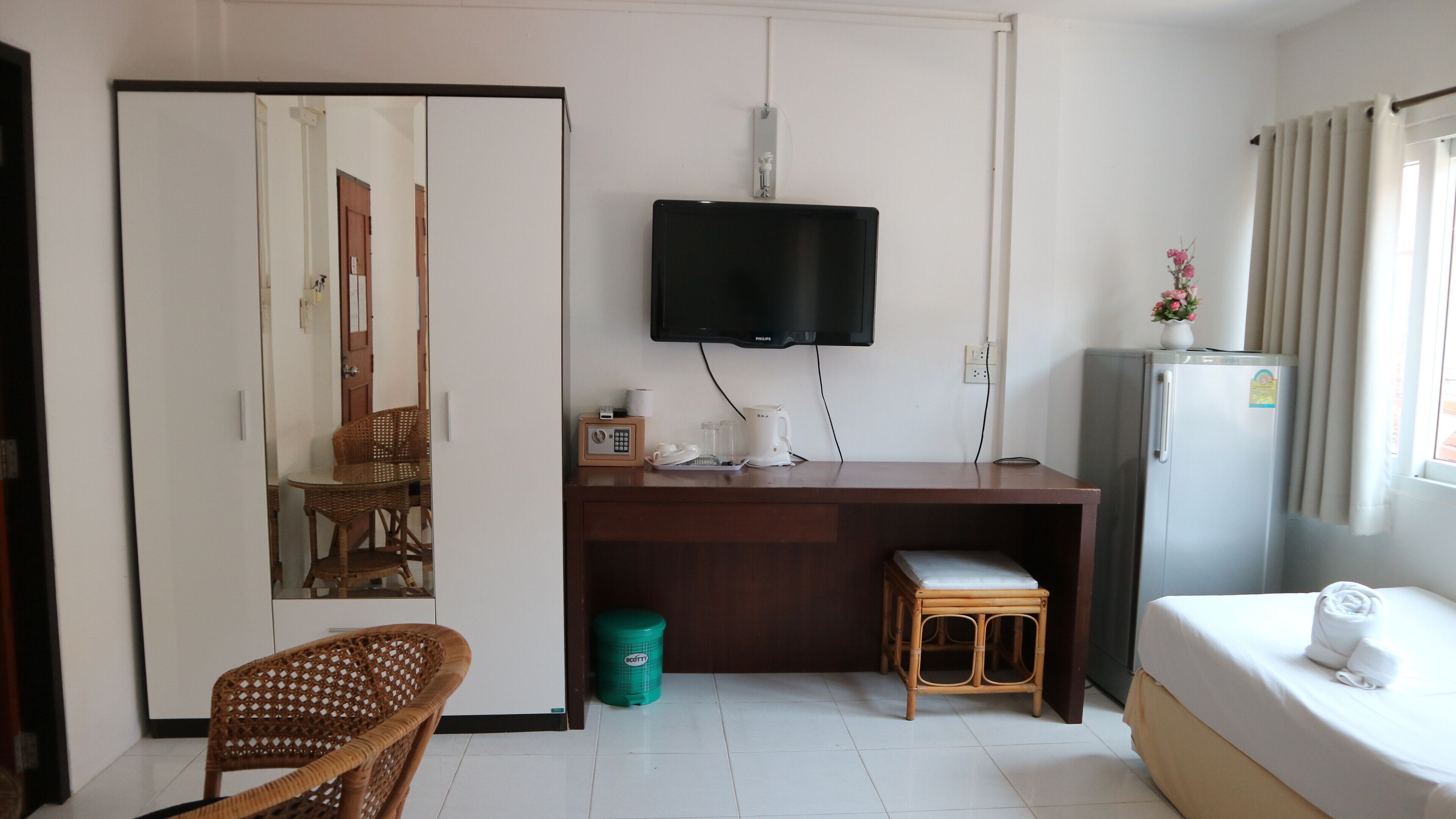 A hotel room with a bed, a desk with a television, a chair, a small stool, a trash bin, and a refrigerator.