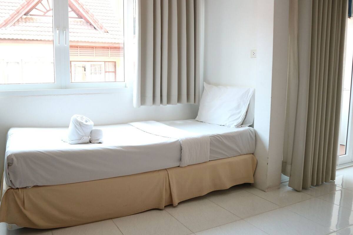 Ein Einzelbett mit weißer Matratze und hellbeiger Bettdecke, einem weißen Kopfkissen und einem zusammengerollten Handtuch darauf.