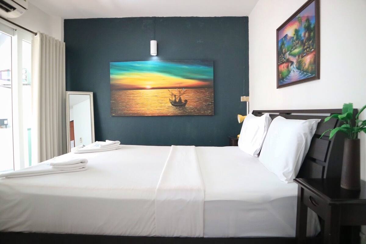 Habitación de hotel con una cama grande, un cuadro de un atardecer sobre el agua, una lámpara de noche y una planta en maceta.