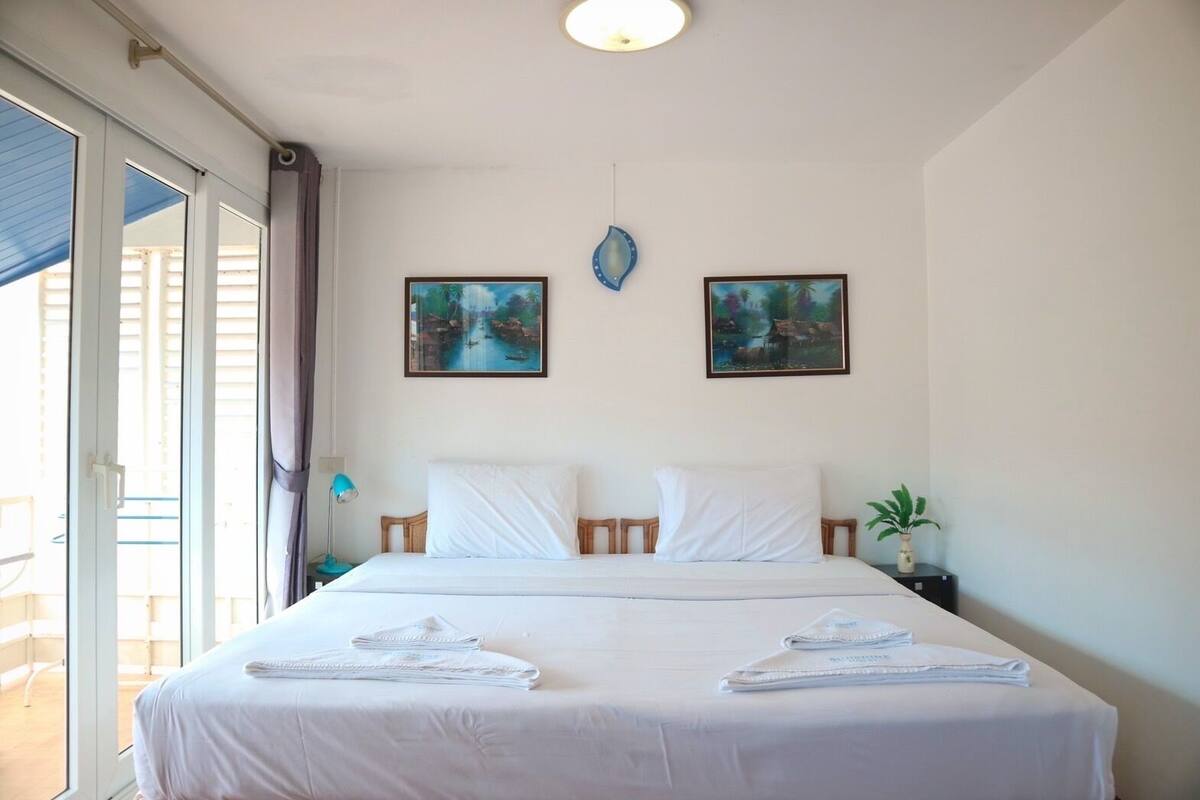 Habitación de hotel con una cama, dos cuadros, una mesita de noche, una lámpara y una planta en maceta.