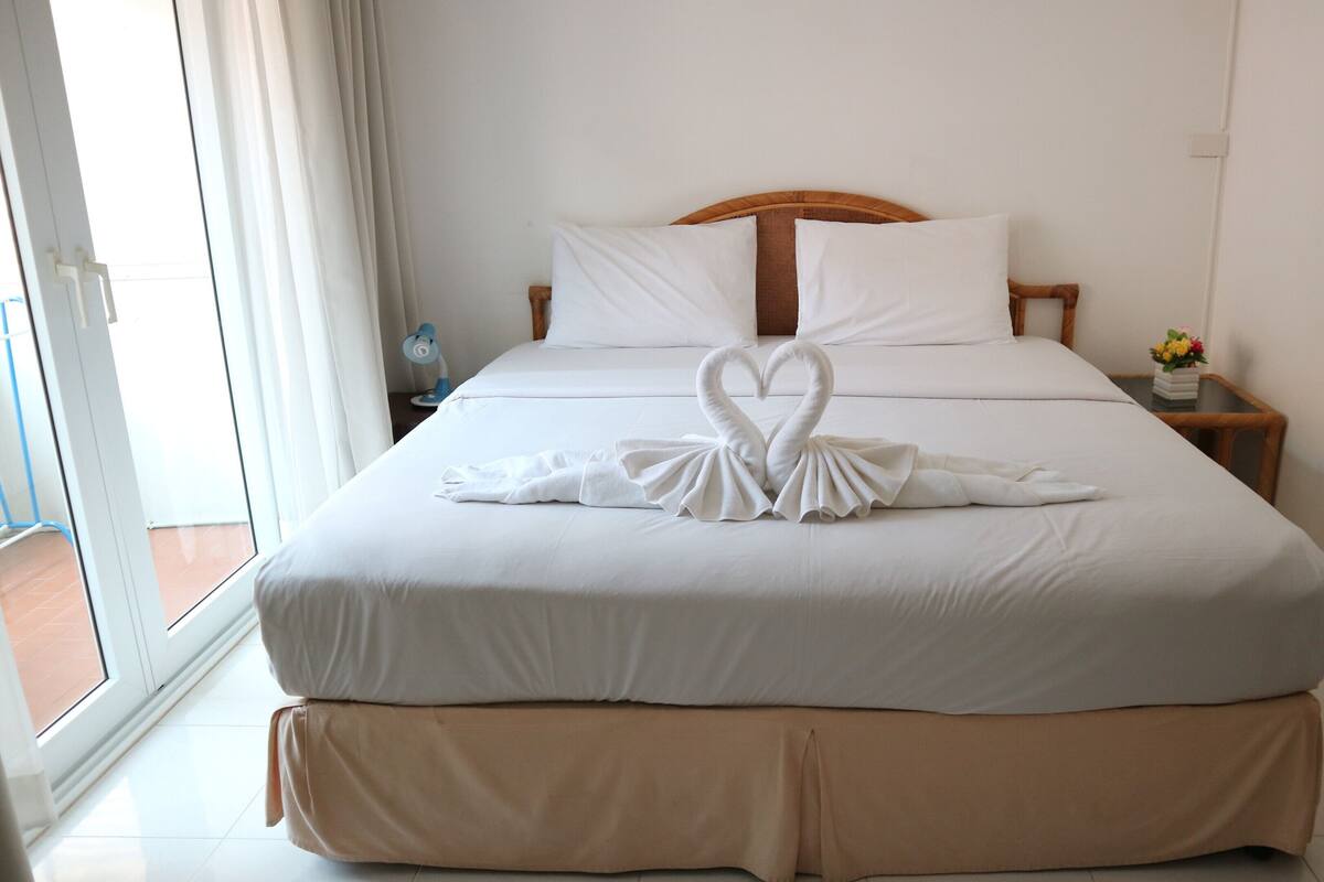 Habitación de hotel con una cama grande, cabecera de madera, ropa de cama blanca y decoración de toalla con forma de cisne.