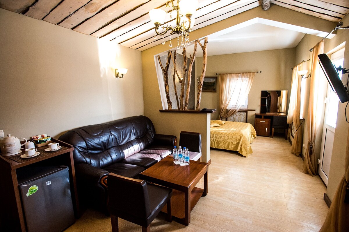 Una habitación acogedora con un sofá de cuero, una mesa de madera con botellas de agua, un refrigerador pequeño, una cama con colcha amarilla y un espejo con diseño de rama de árbol.