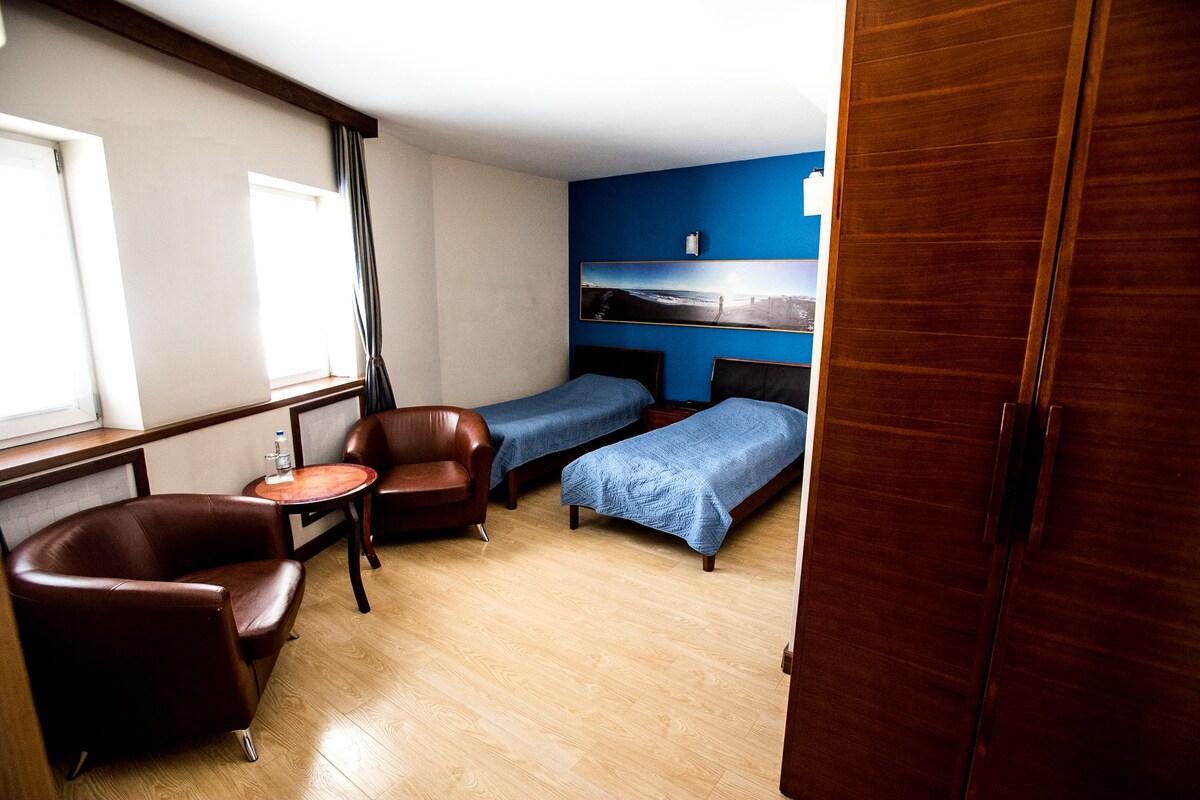 Una habitación con suelo de madera, un sillón de cuero marrón, una mesita redonda pequeña, dos camas con ropa de cama azul y un armario de madera.