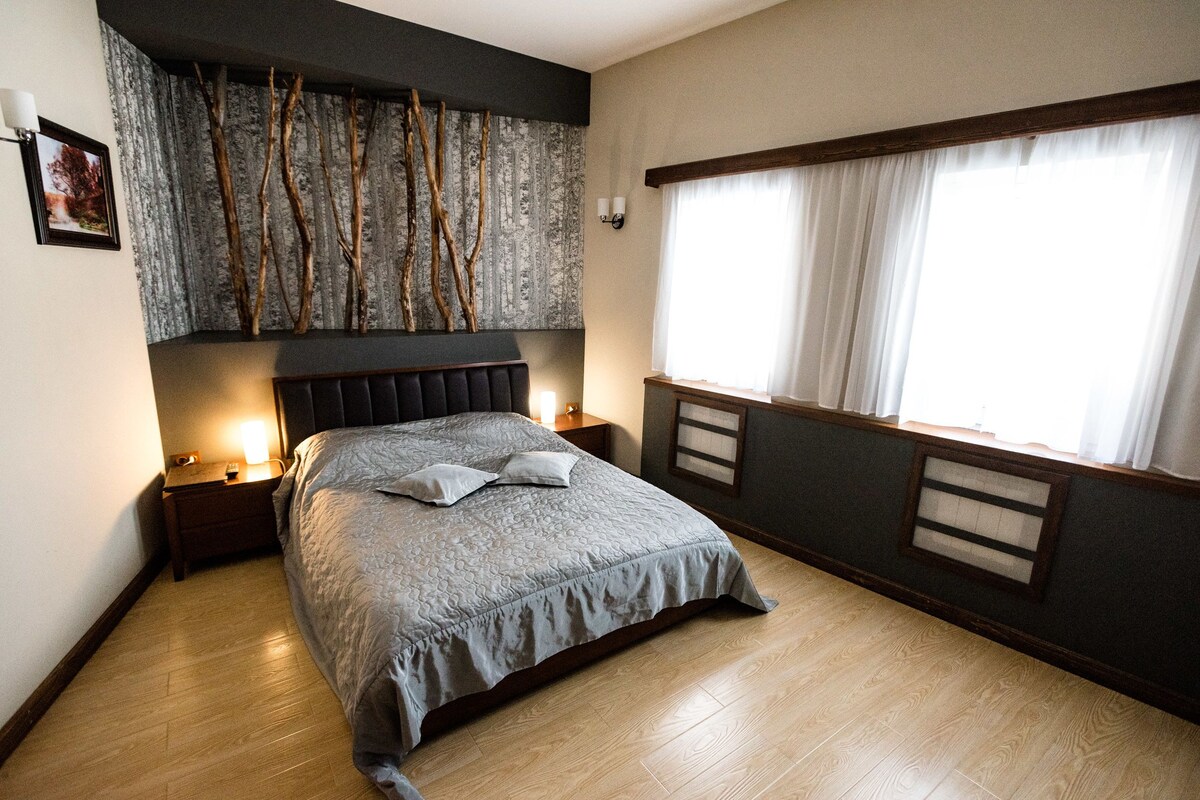 Un dormitorio ordenado con una cama grande, una mesita de noche, una lámpara de pared y un ventanal con cortinas.