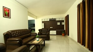 A modern living room with a brown leather sofa, a matching armchair, and a coffee table with a plant. A kitchen is visible in the background with wooden cabinets and a countertop. - Fresh Living Prime Hi Tech (Hyderabad)