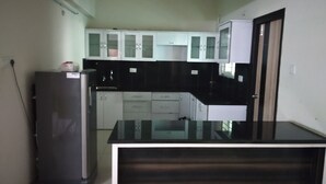 A compact kitchen with white cabinets, a black countertop, and a refrigerator. - Fresh Living Prime Hi Tech (Hyderabad)