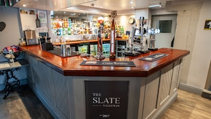 A bar area with a polished wooden counter, a variety of bottles, and a clock on the wall. - The Slate (Bangor)