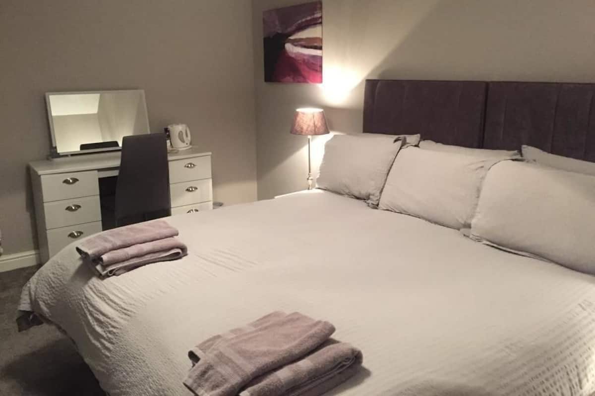 A neatly made bed with pillows and folded towels, a bedside lamp, a desk with a mirror, and a wall-mounted artwork.