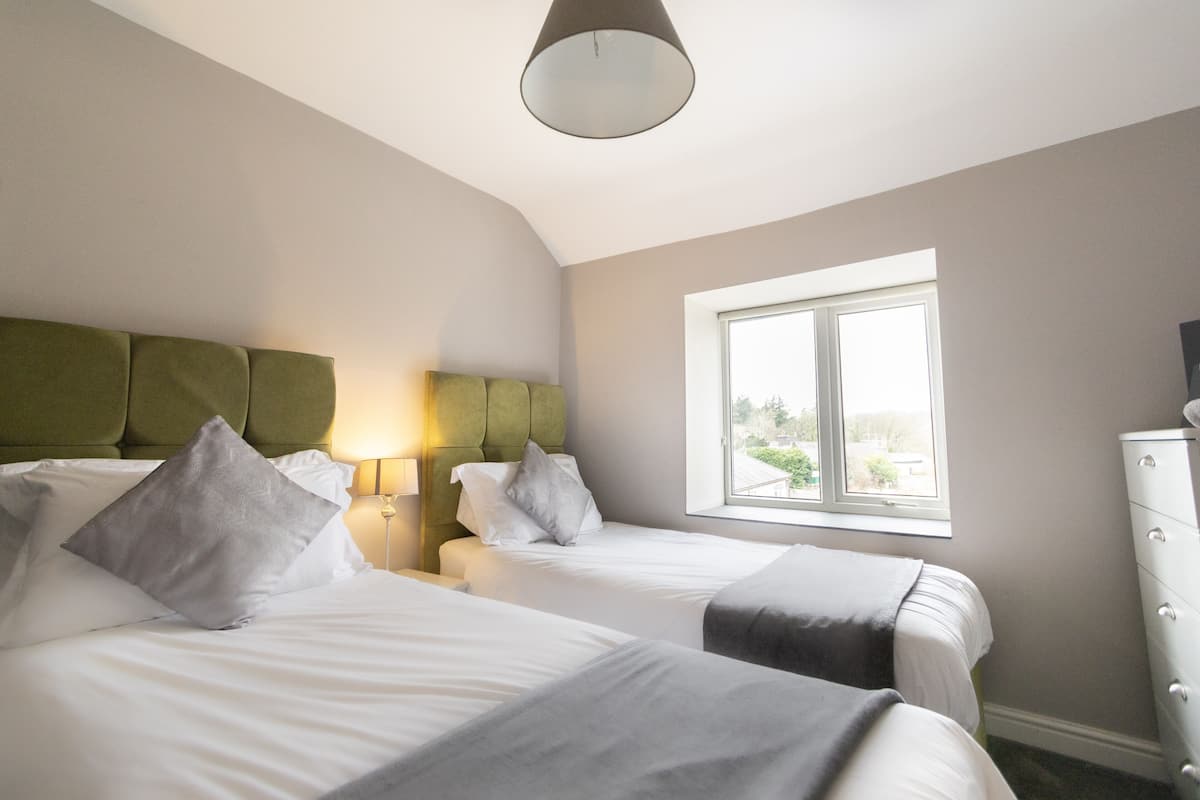 A hotel room with two beds, a green headboard, a window with a view, and a white dresser.