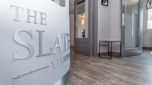 A reception desk with the sign "THE SLATE Y LLECHRA" and a modern interior with a wooden floor and glass doors. - The Slate (Bangor)