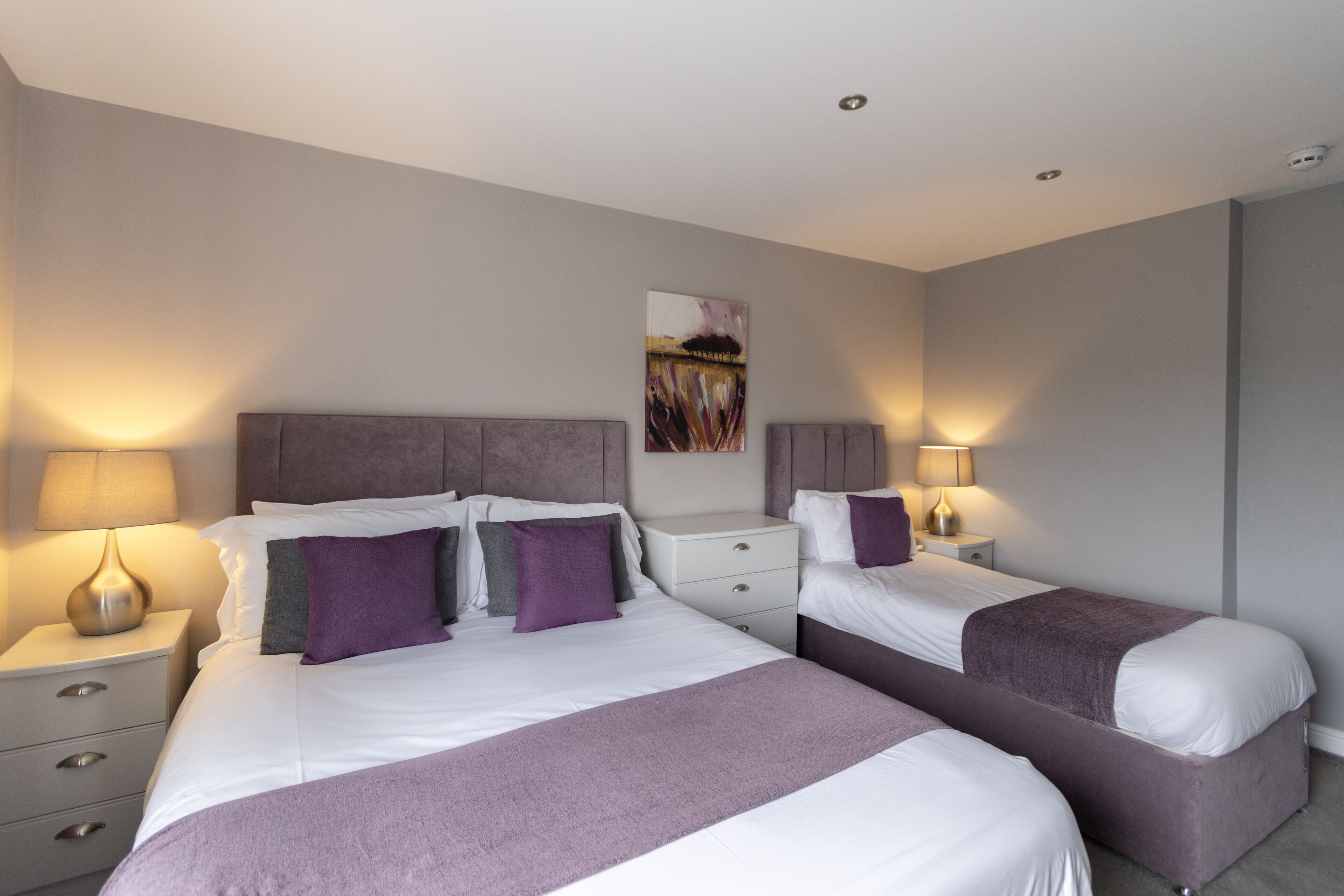 A modern bedroom with two beds, a headboard, bedside tables, and wall-mounted lamps.