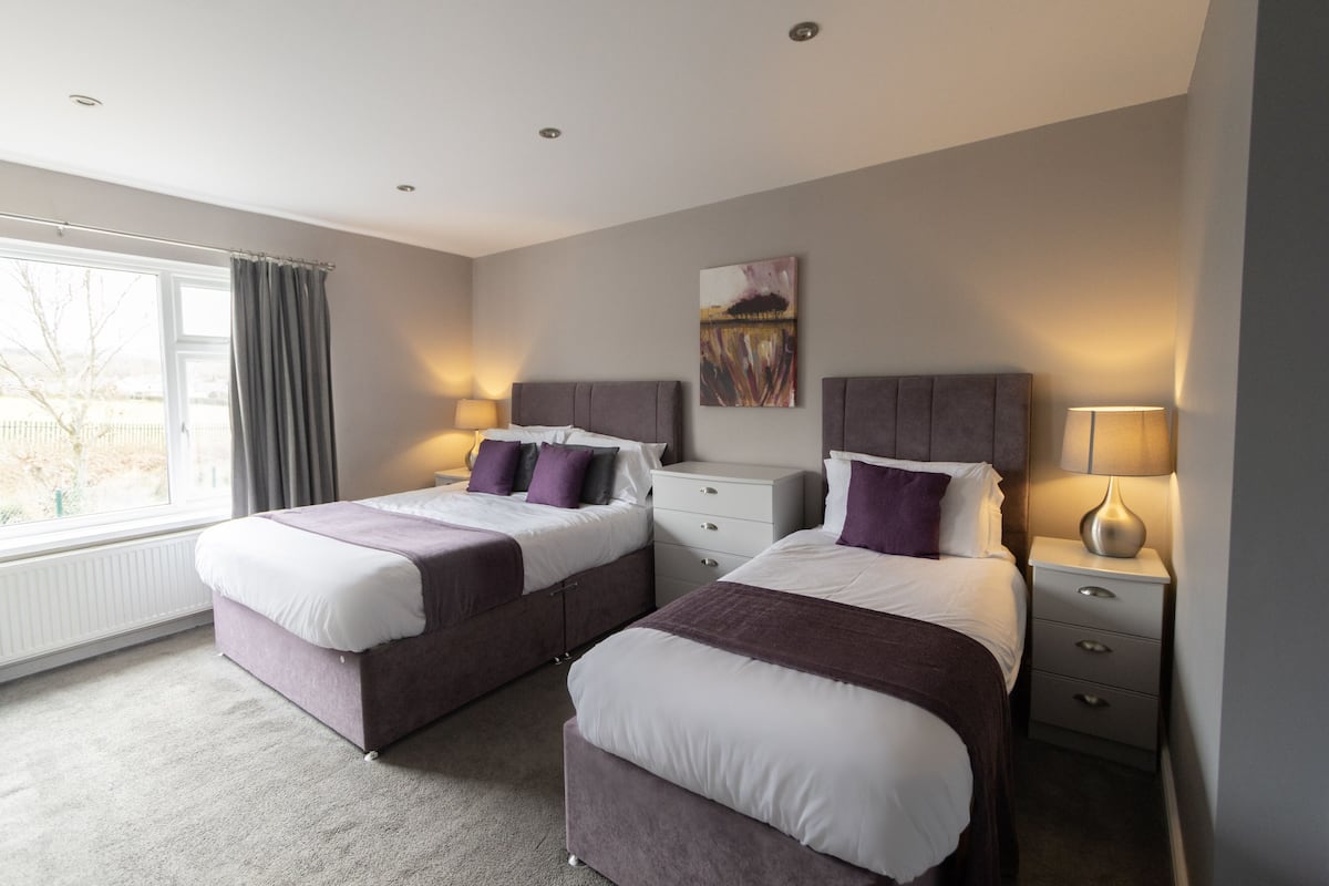 A hotel room with two beds, a window with curtains, a painting on the wall, and a bedside table with a lamp.