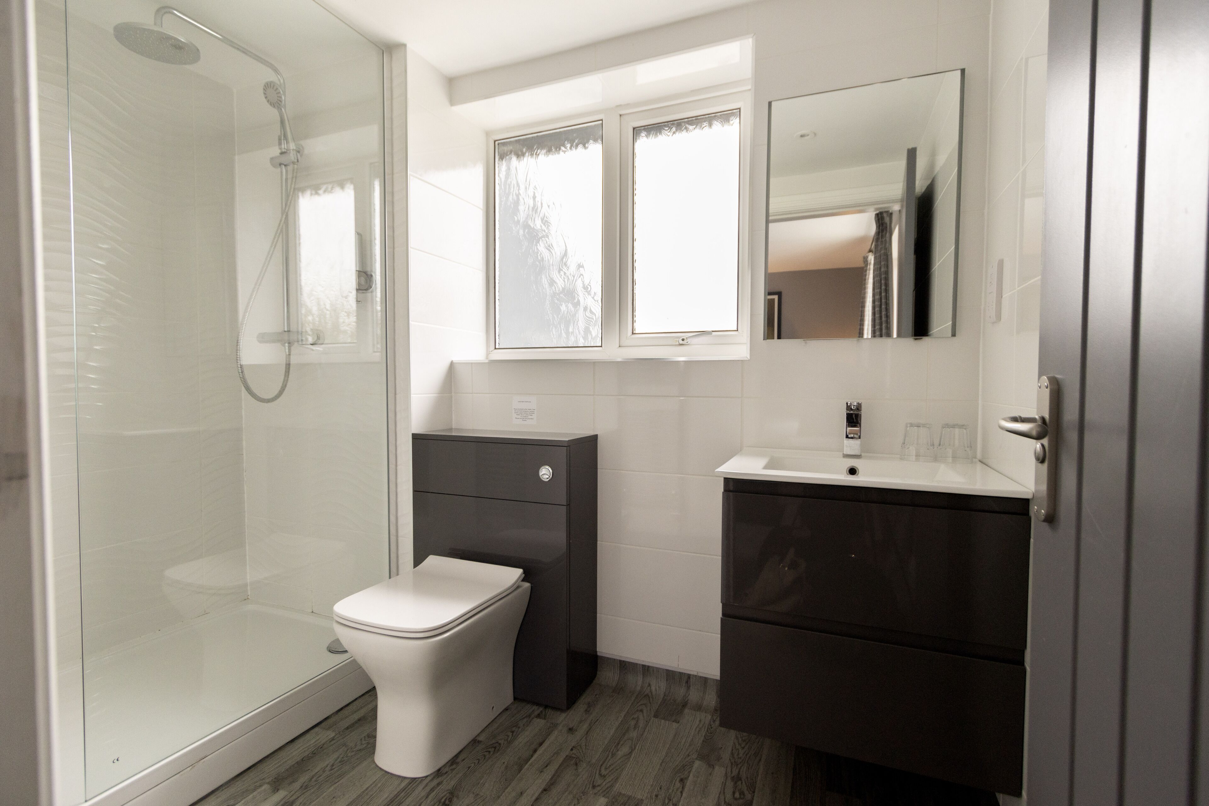 Une salle de bain moderne avec une douche à parois de verre, des toilettes blanches, un meuble-lavabo foncé avec un miroir et une fenêtre au-dessus du lavabo.