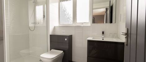 A modern bathroom with a glass-enclosed shower, a white toilet, a dark vanity with a mirror, and a window above the sink.