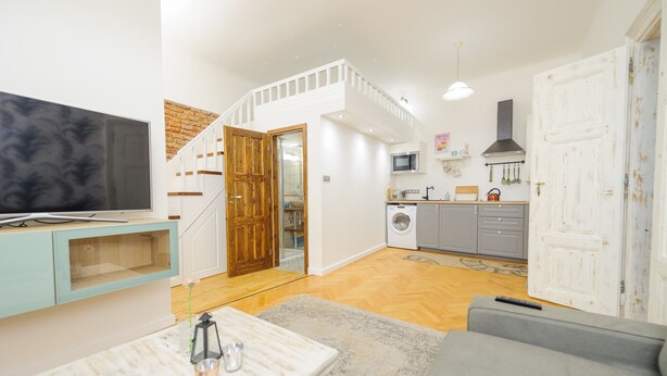 A modern living room with a flat-screen TV, a grey sofa, a coffee table with decorative items, and a staircase leading to a kitchen area with a microwave and washing machine. - Pinkova Apartments (Prague)