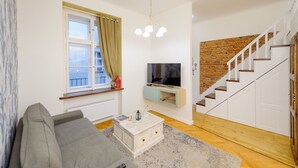 A living room with a grey sofa, a TV on a wooden TV stand, a staircase with a brick wall, and a window with curtains. - Pinkova Apartments (Prague)