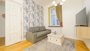 A living room with a grey sofa, a white coffee table, a TV on a wooden cabinet, a patterned wallpaper, and a window with curtains. - Pinkova Apartments (Prague)