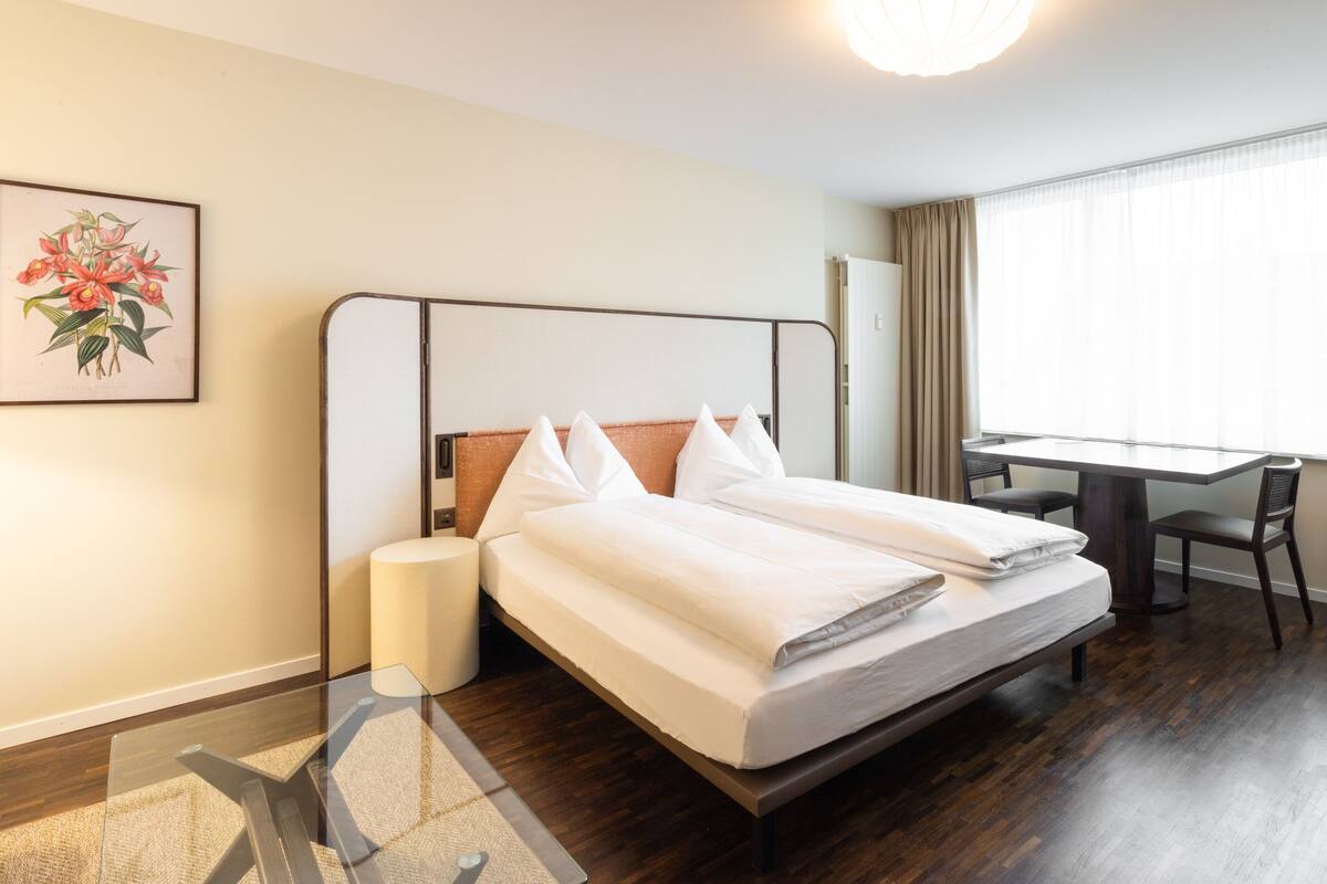 Une chambre d’hôtel comprenant un lit, une table de chevet, une lampe, un bureau, une chaise et une fenêtre avec des rideaux.