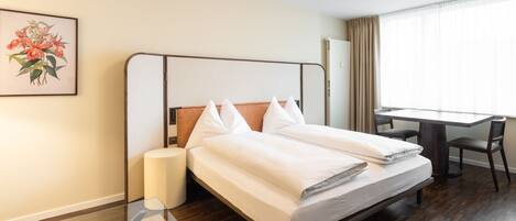 Una habitación de hotel con cama, mesita de noche, lámpara, escritorio, silla y ventana con cortinas.