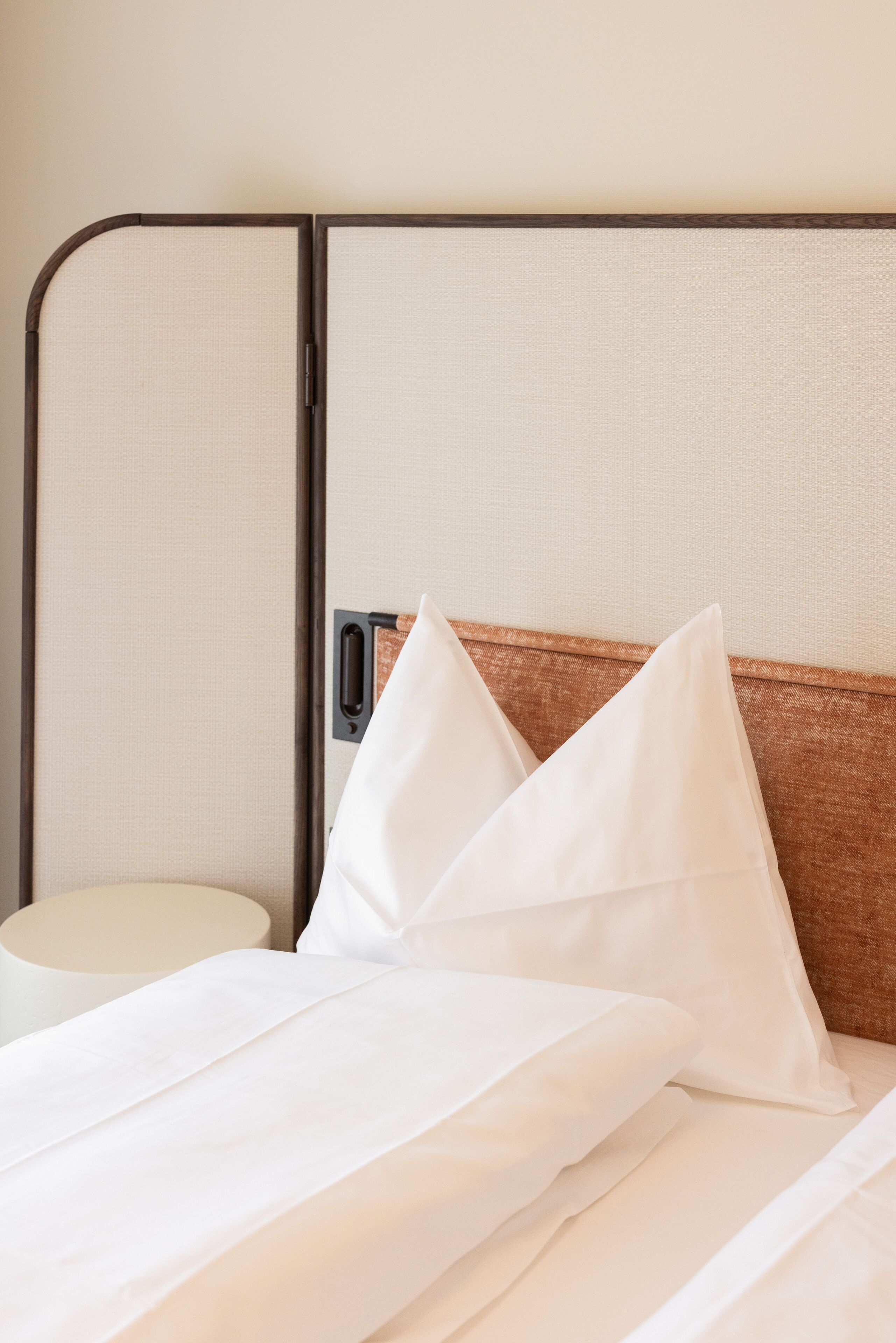 A neatly made bed with white linens and pillows, a bedside table with a lamp, and a partition with a handle.
